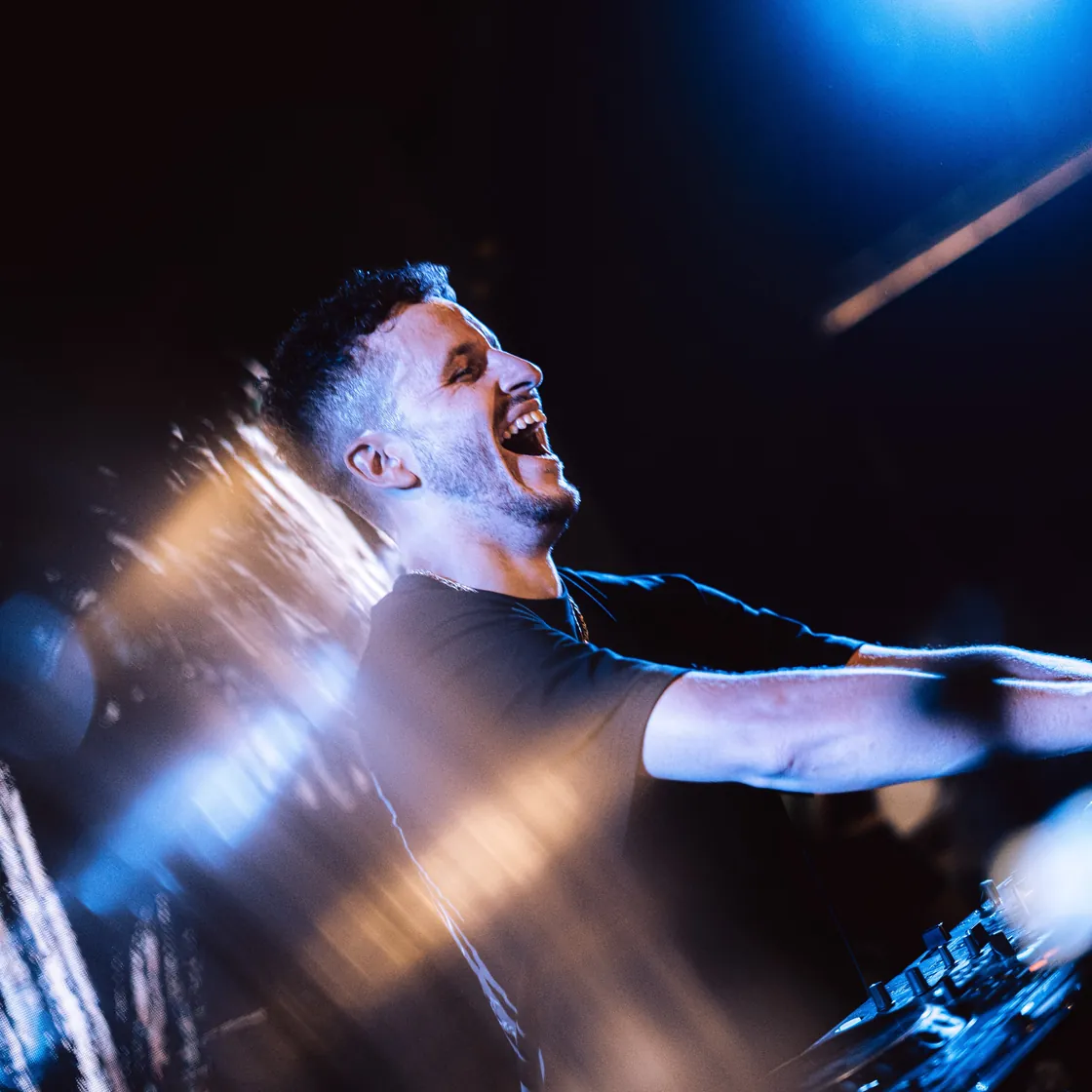 Quyver behind the decks, arms wide under a blue spotlight